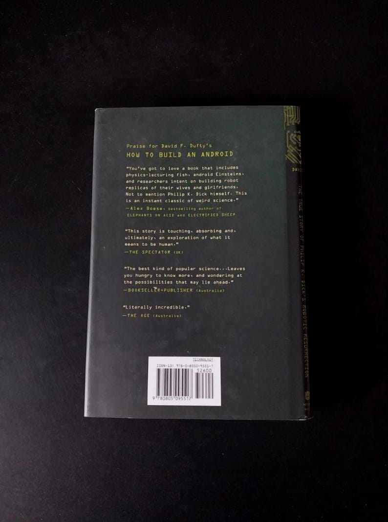 以下が含まれることがあります： David F. Dufty著「How to Build an Android」というタイトルの本の裏表紙。暗い色の表紙には、引用文やバーコードを含む白い文字が書かれています。この本は科学とロボット工学をテーマにしています。