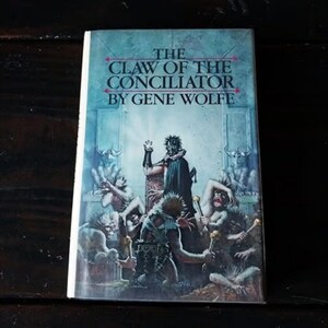 May include: The cover of a paperback book titled "The Claw of the Conciliator" by Gene Wolfe. The cover art depicts a masked figure in a dark robe seated on a throne, surrounded by other figures.