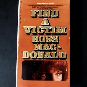 Può includere: Un libro tascabile vintage intitolato "Find a Victim" di Ross Macdonald. La copertina è prevalentemente arancione con scritte dorate in grassetto. Il libro presenta gli occhi di una donna in un rettangolo scuro in basso. Il testo recita "A Lew Archer Novel."