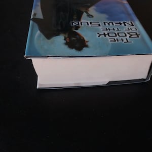Puede incluir: Un libro de tapa dura titulado "The Book of the New Sun" con una cubierta azul y blanca. La cubierta presenta una ilustraci&oacute;n de una figura contra un cielo iluminado por la luna. El lomo del libro es visible, con p&aacute;ginas blancas.