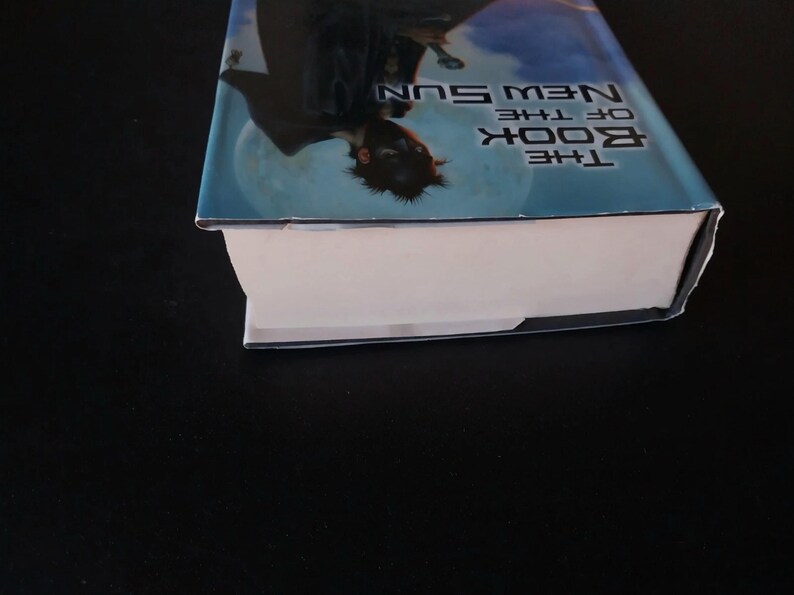 Puede incluir: Un libro de tapa dura titulado "The Book of the New Sun" con una cubierta azul y blanca. La cubierta presenta una ilustraci&oacute;n de una figura contra un cielo iluminado por la luna. El lomo del libro es visible, con p&aacute;ginas blancas.