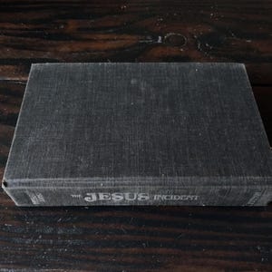 以下が含まれることがあります： 暗い木製の表面にある「The Jesus Incident」というタイトルの濃い灰色の本。本はテクスチャ加工された表紙で、タイトルは明るい灰色で印刷されています。本は長方形で、閉じているように見えます。