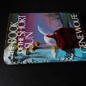 Puede incluir: Un libro titulado "THE BOOK OF THE SHORT SUN" de Gene Wolfe. La portada presenta una ilustraci&oacute;n surrealista con una figura, una luna y estructuras extra&ntilde;as, sobre un cielo azul y morado.