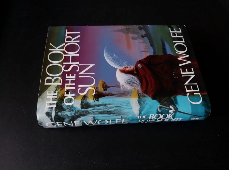 Puede incluir: Un libro titulado "THE BOOK OF THE SHORT SUN" de Gene Wolfe. La portada presenta una ilustraci&oacute;n surrealista con una figura, una luna y estructuras extra&ntilde;as, sobre un cielo azul y morado.