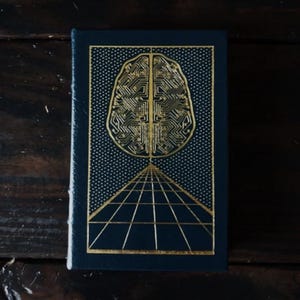 Puede incluir: Un libro azul oscuro con un diseño de cerebro dorado en la portada. El diseño del cerebro está hecho de patrones de circuitos. Debajo del cerebro hay un diseño de cuadrícula dorada. La portada tiene un patrón de puntos.