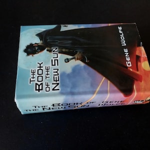 Puede incluir: Un libro de bolsillo titulado "The Book of the New Sun" de Gene Wolfe. La portada presenta una figura con capa negra y espada sobre un fondo azul y naranja. El libro est&aacute; sobre una superficie negra.