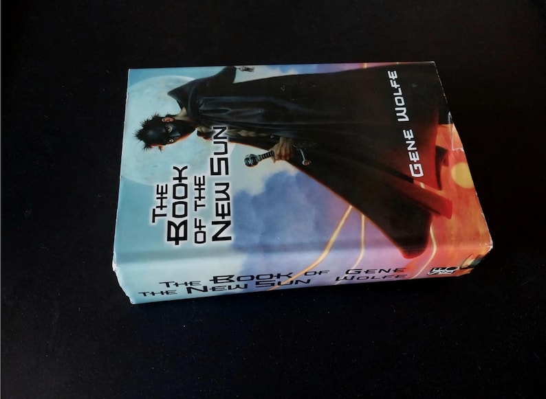 Puede incluir: Un libro de bolsillo titulado "The Book of the New Sun" de Gene Wolfe. La portada presenta una figura con capa negra y espada sobre un fondo azul y naranja. El libro est&aacute; sobre una superficie negra.