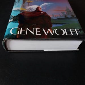 Puede incluir: Un libro de tapa dura con una cubierta colorida que presenta una figura con una capa marr&oacute;n sobre un fondo de un planeta y cielo. El nombre del autor, GENE WOLFE, se muestra de forma destacada.