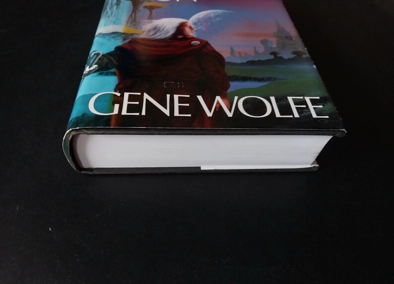 Puede incluir: Un libro de tapa dura con una cubierta colorida que presenta una figura con una capa marr&oacute;n sobre un fondo de un planeta y cielo. El nombre del autor, GENE WOLFE, se muestra de forma destacada.