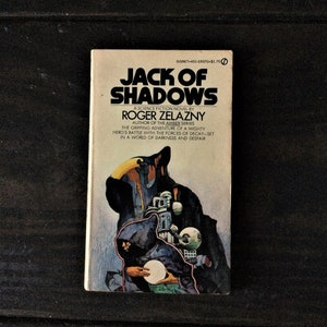 May include: A paperback book cover with a dark blue background and a shadowy figure in a hooded cloak. The title of the book is "Jack of Shadows" by Roger Zelazny. The book is a science fiction novel and part of the Amber series.