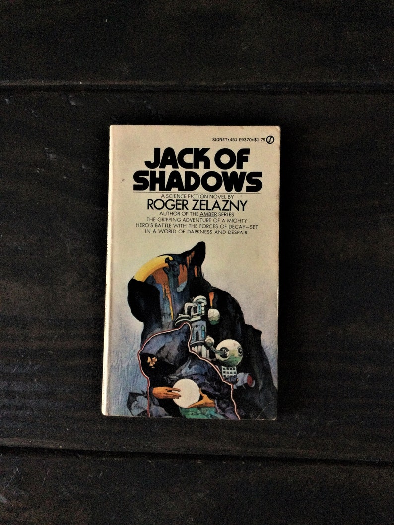 May include: A paperback book cover with a dark blue background and a shadowy figure in a hooded cloak. The title of the book is "Jack of Shadows" by Roger Zelazny. The book is a science fiction novel and part of the Amber series.