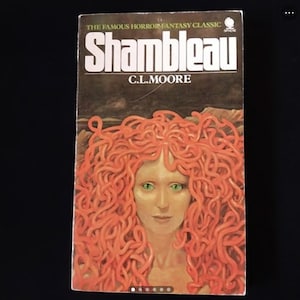 Op de afbeelding: Een paperback boekomslag met de titel "Shambleau" in witte letters op een bruine achtergrond. De omslag toont een vrouw met rood haar gemaakt van slangen en groene ogen.