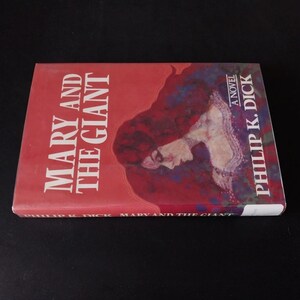 以下が含まれることがあります： フィリップ・K・ディック著の小説「メアリー・アンド・ザ・ジャイアント」。本の表紙は赤色を基調とし、タイトルは大きな白い文字で書かれています。赤い髪の女性のイラストが描かれています。