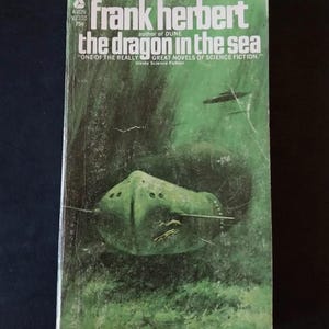 May include: Vintage paperback book cover for "The Dragon in the Sea" by Frank Herbert. The cover features a green submarine illustration with a dark green underwater background. The title and author's name are in white and green text.