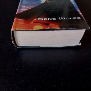 Puede incluir: Un libro de tapa dura con una sobrecubierta que presenta una escena de puesta de sol con una silueta oscura. El nombre del autor, Gene Wolfe, est&aacute; impreso en blanco. Las p&aacute;ginas del libro son blancas y la cubierta est&aacute; ligeramente desgastada.