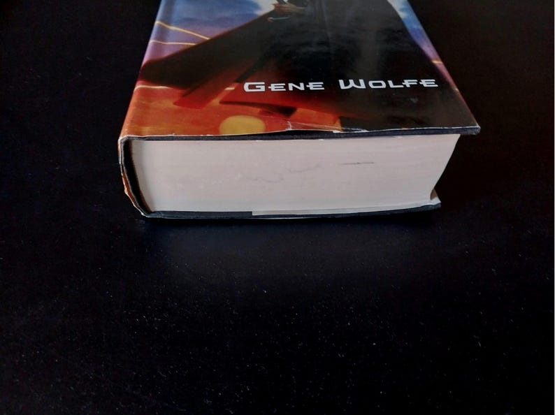 Puede incluir: Un libro de tapa dura con una sobrecubierta que presenta una escena de puesta de sol con una silueta oscura. El nombre del autor, Gene Wolfe, est&aacute; impreso en blanco. Las p&aacute;ginas del libro son blancas y la cubierta est&aacute; ligeramente desgastada.