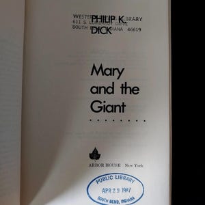 以下が含まれることがあります： 黒字で「Mary and the Giant」と書かれた本のページ。著者名、フィリップ・K・ディックも表示されています。1987年4月29日付けのインディアナ州サウスベンドの図書館スタンプも表示されています。