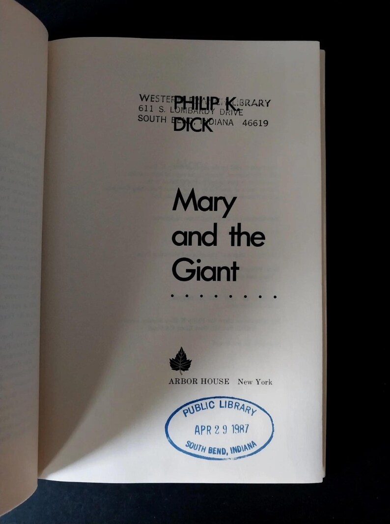 以下が含まれることがあります： 黒字で「Mary and the Giant」と書かれた本のページ。著者名、フィリップ・K・ディックも表示されています。1987年4月29日付けのインディアナ州サウスベンドの図書館スタンプも表示されています。