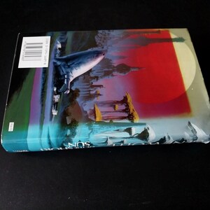 Puede incluir: Un libro de bolsillo con una cubierta colorida que presenta una ciudad futurista, un gran c&iacute;rculo rojo y blanco, y un objeto azul que se asemeja a una nave espacial. El libro est&aacute; sobre una superficie negra.