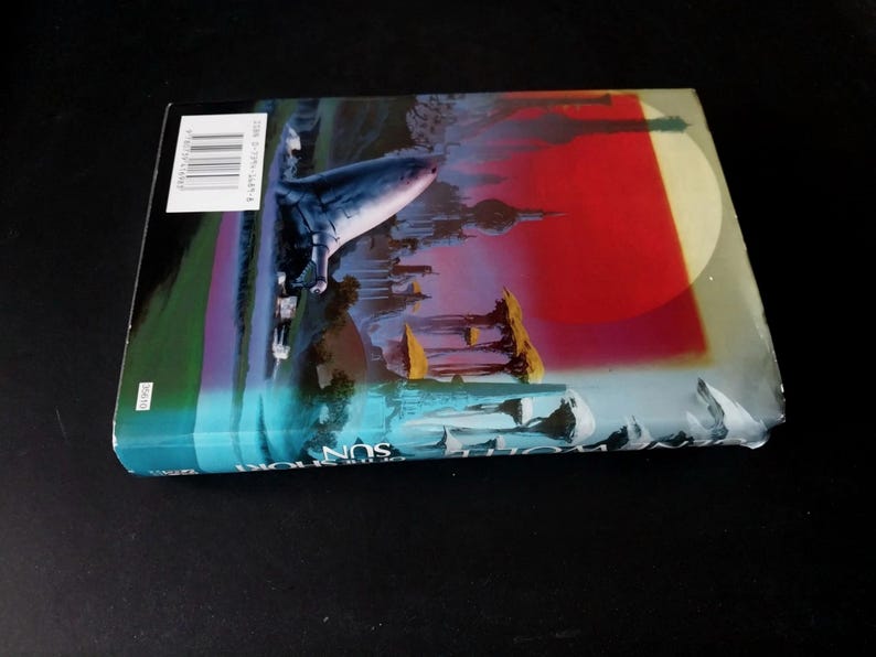 Puede incluir: Un libro de bolsillo con una cubierta colorida que presenta una ciudad futurista, un gran c&iacute;rculo rojo y blanco, y un objeto azul que se asemeja a una nave espacial. El libro est&aacute; sobre una superficie negra.
