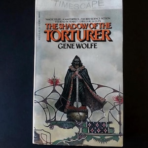 May include: A vintage paperback book titled "The Shadow of the Torturer" by Gene Wolfe. The cover features a cloaked figure holding a sword, set against a backdrop of intricate, fantastical artwork. The book is part of the Timescape series.