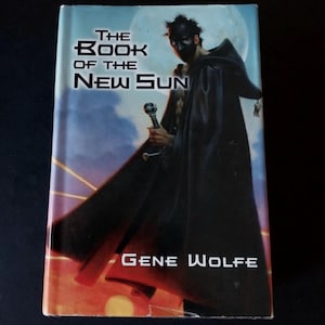 May include: A paperback copy of "The Book of the New Sun" by Gene Wolfe. The cover depicts a cloaked figure with a mask and sword, set against a moonlit backdrop. The book's title and author's name are clearly visible.