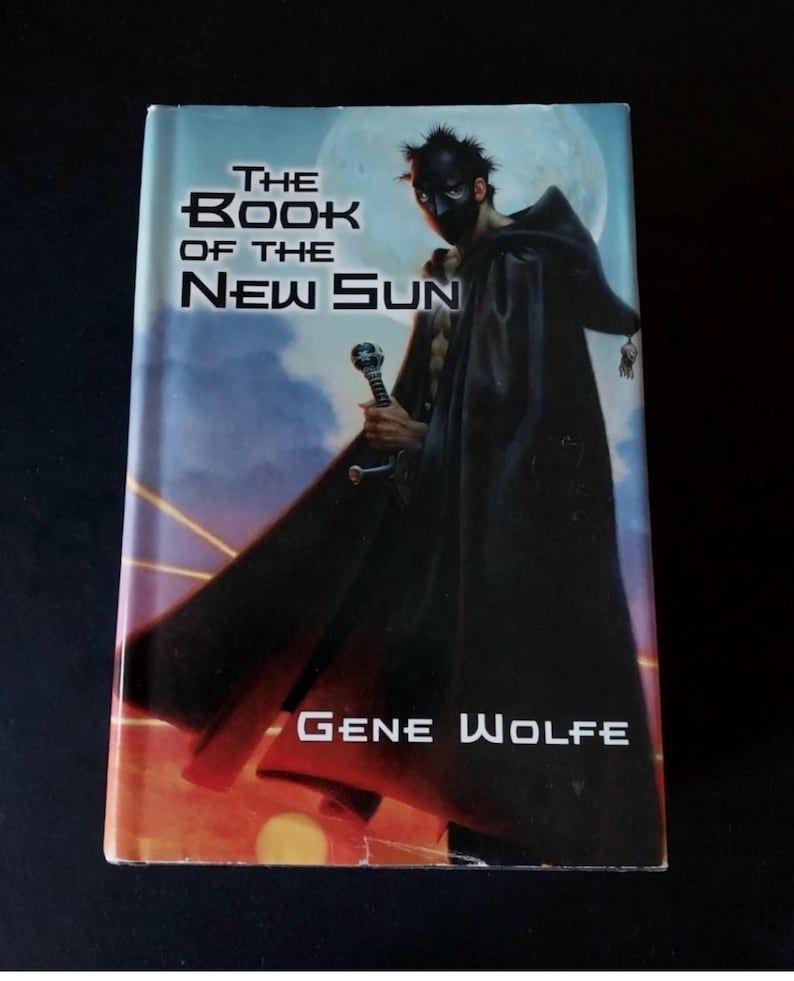 Puede incluir: Un libro de bolsillo titulado "The Book of the New Sun" de Gene Wolfe. La portada muestra una figura enmascarada con una capa negra sosteniendo una espada, sobre un fondo de luna. El t&iacute;tulo y el nombre del autor son visibles.