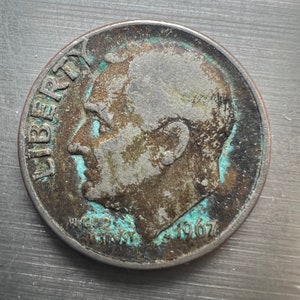 May include: A silver-colored 1967 United States dime coin with a portrait of Franklin D. Roosevelt on the obverse and the inscription "LIBERTY" on the left side. The reverse side features the inscription "IN GOD WE TRUST" and the year "1967".