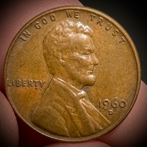 May include: A copper-colored 1960 US penny coin with the profile of Abraham Lincoln on the obverse side. The coin has the inscription "IN GOD WE TRUST" above Lincoln's head and "LIBERTY" below his bust. The date "1960" and the mint mark "D" are visible on the right side of the coin.