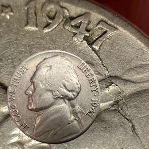 May include: A silver 1947 Jefferson nickel with the profile of Thomas Jefferson. The coin is in focus, with the words "IN GOD WE TRUST" and "LIBERTY" visible. The year "1947" is stamped on the coin.