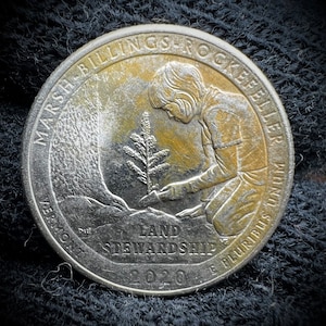 May include: A silver quarter dollar coin with a depiction of a person planting a tree. The coin is from the "Marsh Billings Rockefeller National Memorial" and features the text "Land Stewardship" and the year "2020".