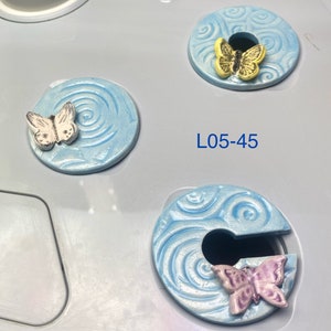 May include: Three ceramic bottle stoppers with a blue and white swirl design. Each stopper features a different colored butterfly: white, yellow, and purple.