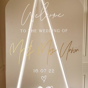 May include: A clear acrylic wedding welcome sign with gold lettering. The sign reads "Welcome to the wedding of Mr. & Mrs. Unwin 16 07 22".