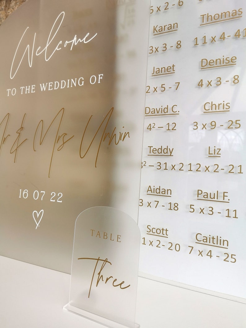 May include: A white acrylic sign with gold lettering that says "Welcome to the Wedding of Mr. & Mrs. Unwin" with the date "16 07 22" and a heart symbol. The sign also has a seating chart with names and table numbers. A small clear acrylic table number sign with gold lettering that says "Table Three" is in the foreground.