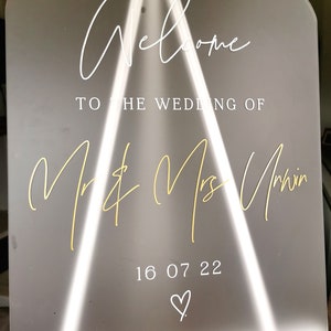May include: A white acrylic wedding welcome sign with gold lettering. The sign reads "Welcome to the wedding of Mr. & Mrs. Unwin 16 07 22" with a heart symbol.