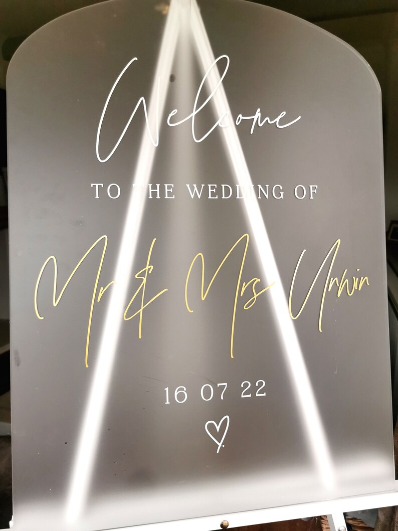 May include: A white acrylic wedding welcome sign with gold lettering. The sign reads "Welcome to the wedding of Mr. & Mrs. Unwin 16 07 22" with a heart symbol.