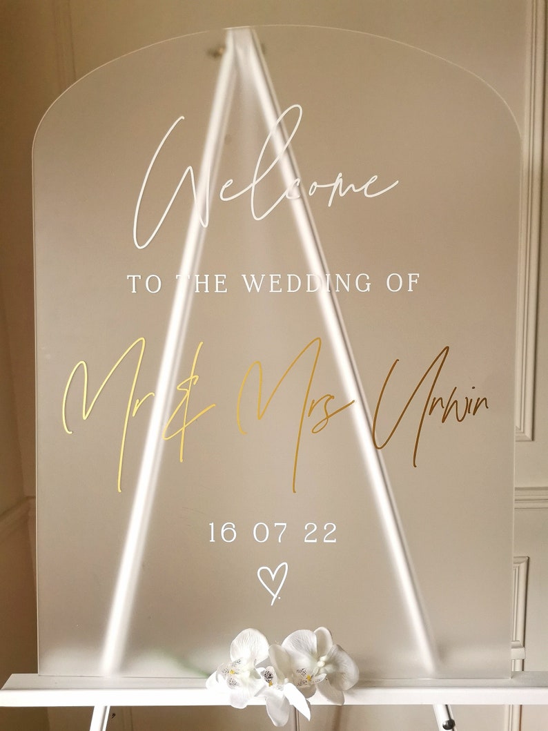May include: A clear acrylic wedding welcome sign with gold lettering. The sign reads "Welcome to the wedding of Mr. & Mrs. Unwin 16 07 22".
