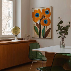 May include: An interior shot featuring a framed floral art piece with orange flowers and blue centers. Two transparent green chairs with metal legs are positioned near a white table. A decorative lamp and wooden cabinet are also visible.