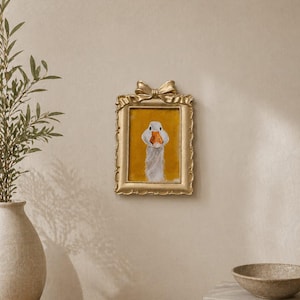 May include: A framed artwork featuring a goose portrait. The painting has a yellow background and a gold frame with a bow at the top. The artwork is hanging on a beige wall.