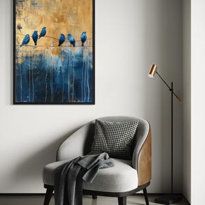 May include: A framed abstract painting features six blue birds perched on a wire against a textured background of gold and blue. The artwork hangs above a gray armchair with a houndstooth pillow and a dark gray blanket.