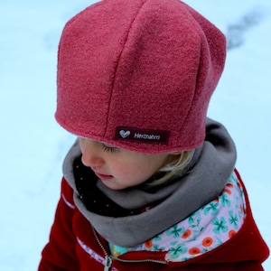 May include: A young child wearing a red hooded jacket with a floral pattern on the inside. The child is wearing a pink wool hat with a white heart and the text "Herznah" on a label. The child is looking down.