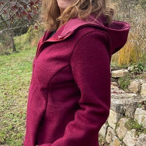Könnte beinhalten: Burgunderfarbene Kapuzenjacke mit durchgehendem Reißverschluss. Die Jacke hat ein kontrastierendes burgunderfarbenes Futter in der Kapuze und eine taillierte Passform. Die Person trägt die Jacke im Freien in einer natürlichen Umgebung.
