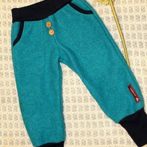 May include: Teal blue children's pants with black waistband and cuffs. The pants have two front pockets and two button closures. The pants have a small brown label with the word "Bambini" on the right leg.