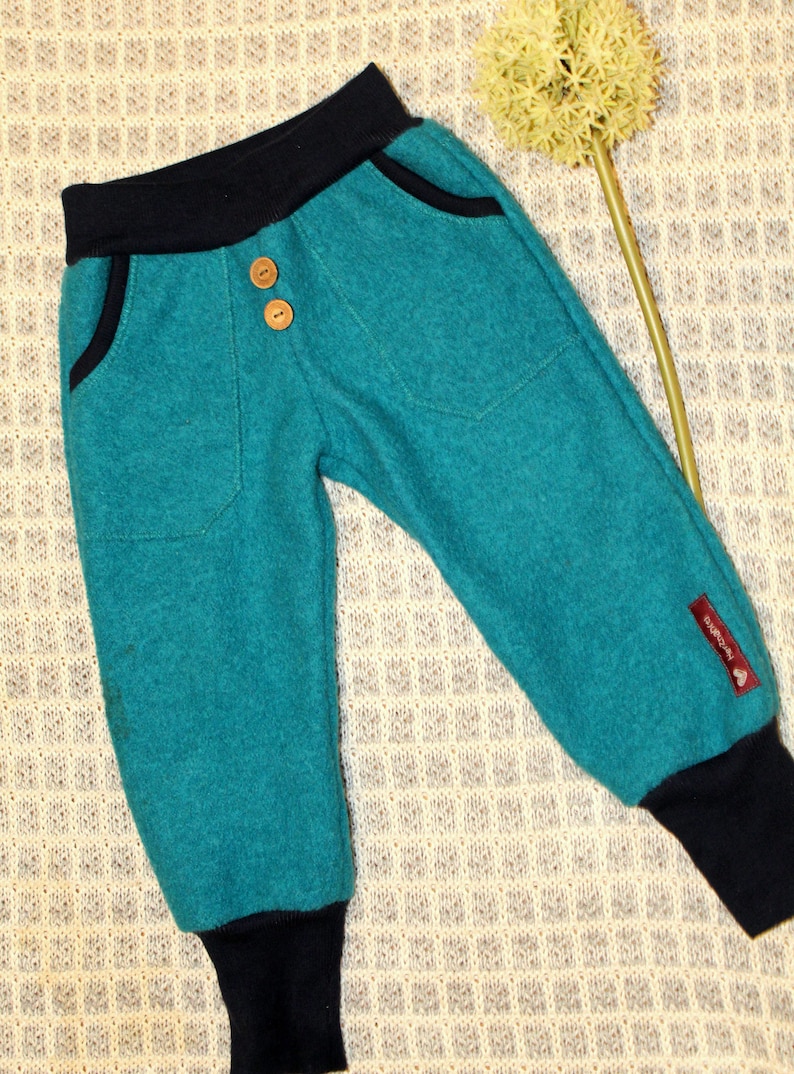 May include: Teal blue children's pants with black waistband and cuffs. The pants have two front pockets and two button closures. The pants have a small brown label with the word "Bambini" on the right leg.