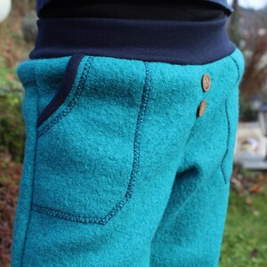May include: Teal blue fleece pants with a dark blue waistband and two brown buttons. The pants have a large pocket on the front.