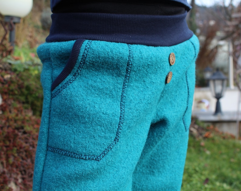 May include: Teal blue fleece pants with a dark blue waistband and two brown buttons. The pants have a large pocket on the front.