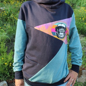 Könnte beinhalten: Ein farbblockierter Hoodie in Marineblau, Türkis und Lila mit einer Grafik eines Affen mit Hut. Der Hoodie hat einen hohen Kragen und lange Ärmel. Die Person trägt eine blaue Jeans.