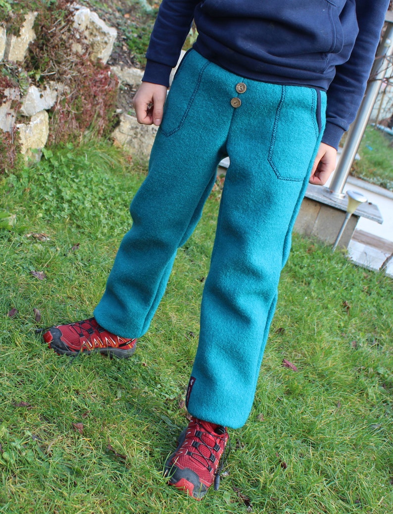 May include: A child wearing teal blue fleece pants with a button fly and side pockets. The pants are worn with red sneakers.