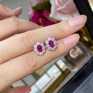 May include: A pair of oval-shaped stud earrings. Each earring features a vibrant, oval-cut ruby center stone surrounded by a halo of small, sparkling white gemstones. The earrings are set in a silver-colored metal.