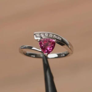 May include: A silver-toned ring featuring a central trillion-cut vibrant pink gemstone held in a prong setting. The band curves around the stone, with a channel-set row of small clear white accent stones embedded along the upper metal arc.